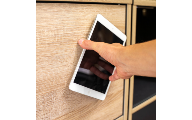 Hand hält ein weißes Tablet mit schwarzem Bildschirm zwischen zwei Schubladen eines hellen Holzschranks
