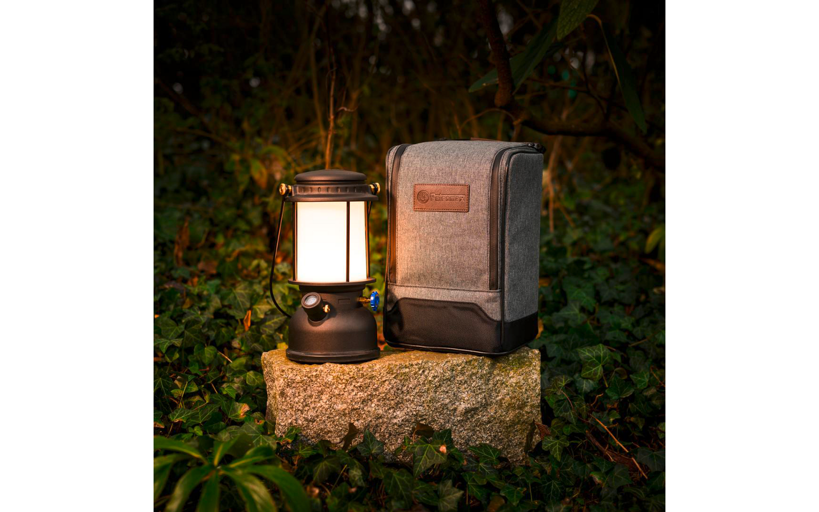 Lanterna accesa con base nera e diffusore bianco accanto a una custodia in tessuto grigia in posizione verticale con cerniera, poste su una pietra rettangolare e circondate da foglie verdi