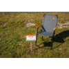 Padded gray folding chair with black metal frame and brown armrests on grass; beside it a small white side table with an orange drinking cup on a book