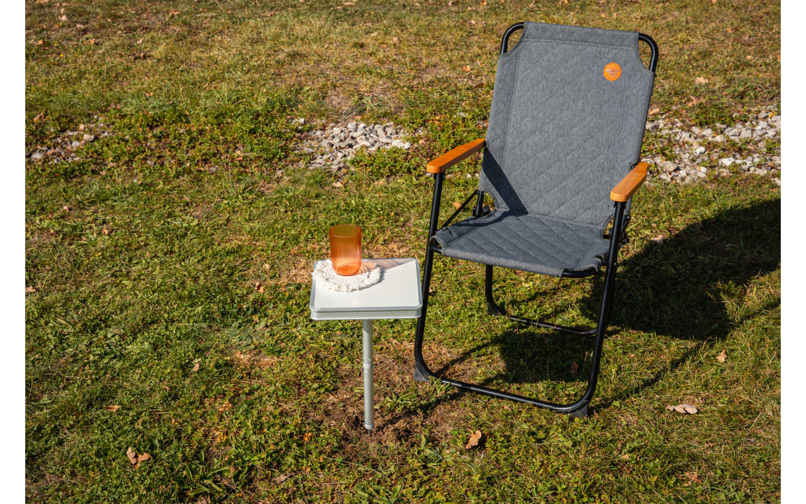 Padded gray folding chair with black metal frame and brown armrests on grass; beside it a small white side table with an orange drinking cup on a book
