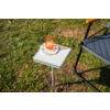 White square side table on grass, with an orange glass tumbler on a white crocheted coaster; a gray chair with a black metal frame visible at the right edge