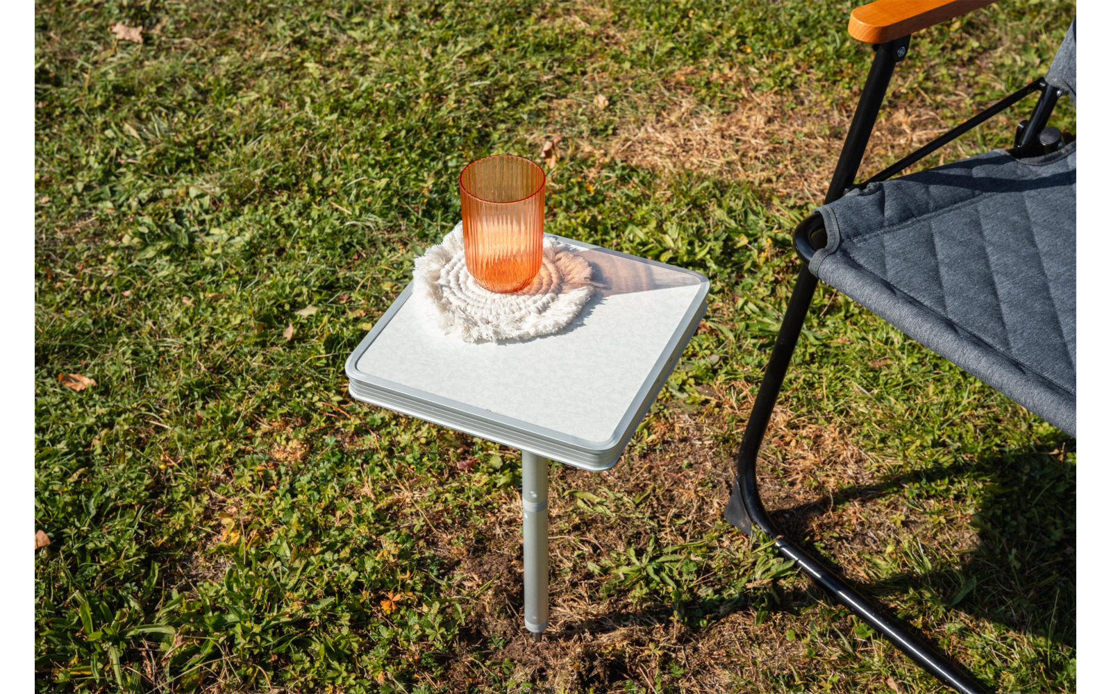 White square side table on grass, with an orange glass tumbler on a white crocheted coaster; a gray chair with a black metal frame visible at the right edge