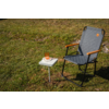 Gray quilted folding chair with wooden armrests and black frame on grass; beside it a small white square side table with a white book and an amber glass