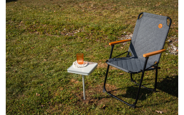 Gray quilted folding chair with wooden armrests and black frame on grass; beside it a small white square side table with a white book and an amber glass