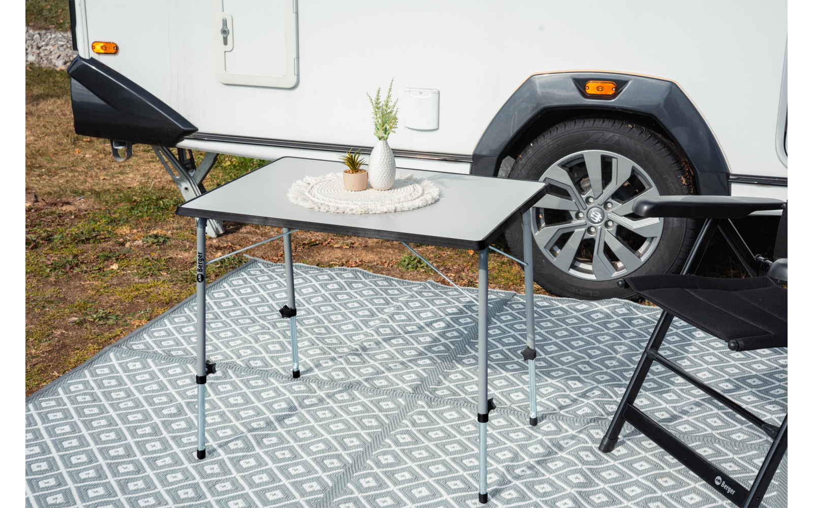 Small folding table with white vase and decorations on a round placemat, two black folding chairs, patterned outdoor mat, and partial view of a white caravan with wheel