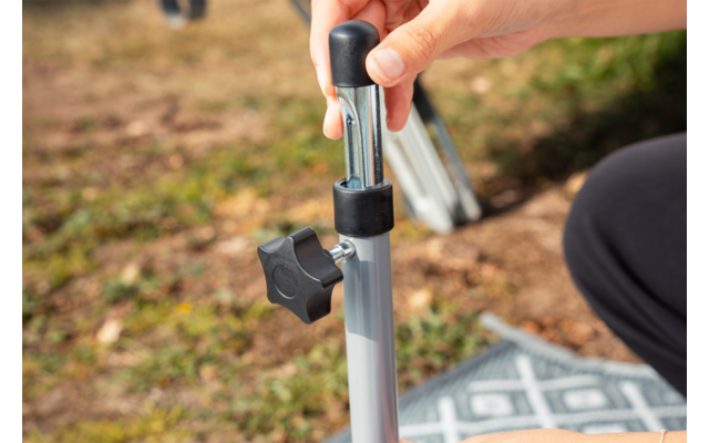Hand holding a silver telescopic pole with a black adjustment knob and locking pin; patterned cloth on grass; dark trouser leg at the image edge
