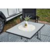 White rectangular camping table with a crocheted doily and small potted plant, black chair behind, patterned gray outdoor rug, grass and part of a caravan wheel visible at left