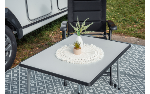 White rectangular camping table with a crocheted doily and small potted plant, black chair behind, patterned gray outdoor rug, grass and part of a caravan wheel visible at left