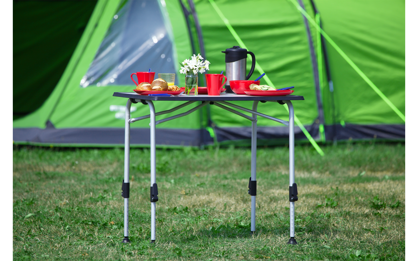 Een inklapbare campingtafel met koffiekopjes, een koffiekruik en een bord voor een groen tent op gras