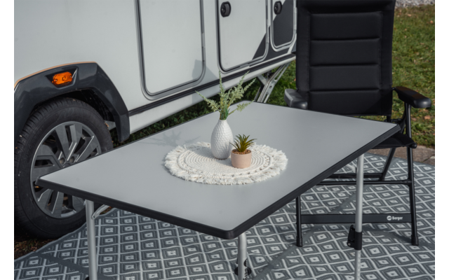 Klapp­tisch vor einem Wohnwagen; auf dem Tisch eine weiße Vase mit Gras und eine kleine Sukkulente auf einem Spitzen­deckchen; schwarzer Campingstuhl daneben; Rad des Wohnwagens und gemusterter Boden­teppich sichtbar