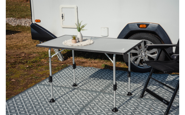 Klapptisch mit zwei kleinen Topfpflanzen und einer Schale mit Muscheln auf einem gemusterten Teppich vor einem weißen Wohnwagen; rechts ein schwarzer Stuhl