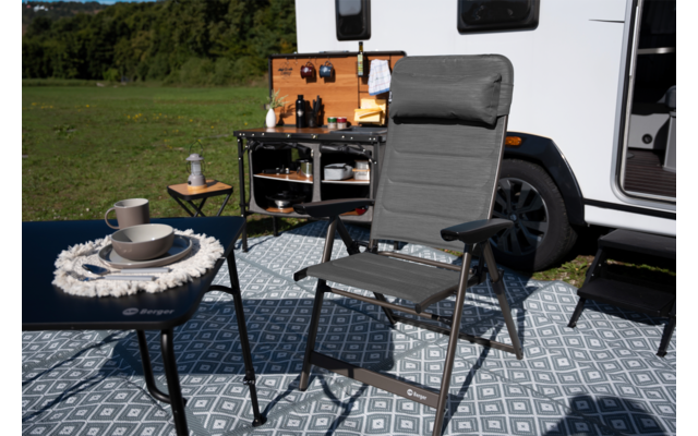 Grijze vouwstoel met armleuningen op een gemusterd vloerkleed voor een witte caravan; twee tafels met kopjes en kommen; geopende buitenkeuken en grasveld met bomen op de achtergrond