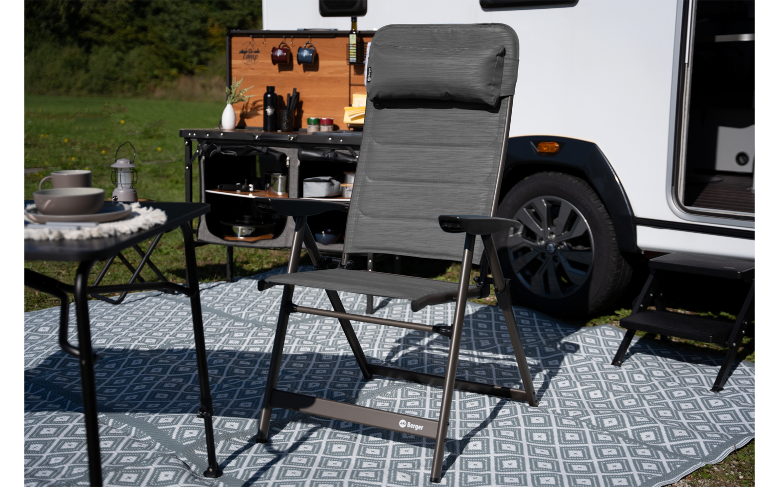 Grijze vouwstoel op een patroon buitenkleed voor een geopend camper, zijkeuken met keukengerei, klein tafeltje met servies, camperwiel en gras op de achtergrond