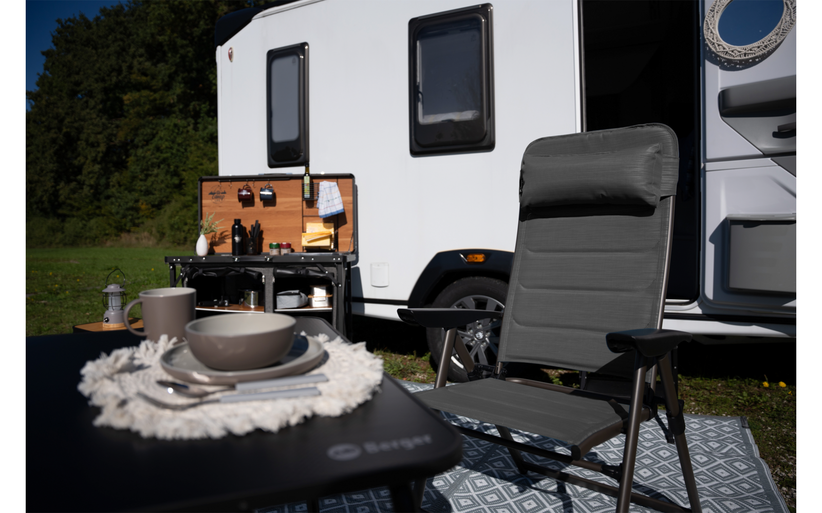 Witte camper op gras; ervoor een grijze campingstoel, klein tafeltje met borden, kom en bestek, een open houten kist met flessen en keukengerei, een geprint grondkleed en bomen op de achtergrond