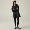 Persona in piedi con le braccia incrociate davanti a uno sfondo chiaro uniforme, indossa un lungo cappotto trapuntato nero, leggings neri, una felpa con cappuccio rosa chiaro e scarpe sportive grigie