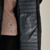 Interno di un cappotto trapuntato blu scuro aperto con tasca interna e due pittogrammi di cura bianchi; a sinistra top rosa chiaro parzialmente visibile e leggings neri, sfondo neutro