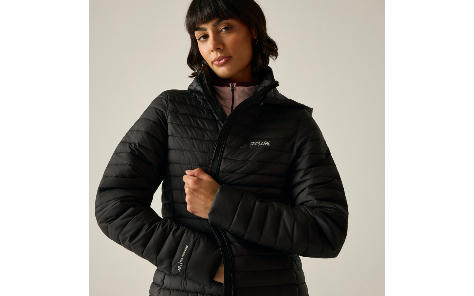 Donna con giacca imbottita nera trapuntata che tiene la cerniera, top rosa chiaro visibile al colletto, sfondo chiaro uniforme