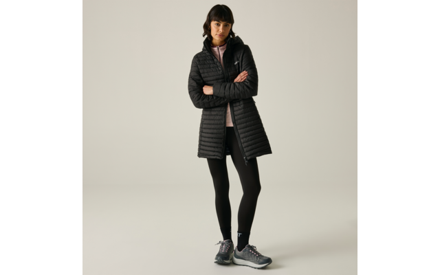 Persona in piedi con le braccia incrociate davanti a uno sfondo chiaro uniforme, indossa un lungo cappotto trapuntato nero, leggings neri, una felpa con cappuccio rosa chiaro e scarpe sportive grigie
