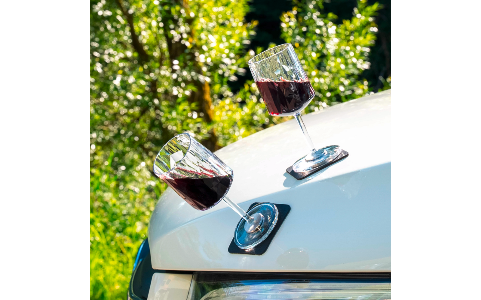 Two glasses of red wine are placed on the hood of a white car
