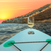 Wine glass on a stand-up paddleboard with a rocky shore and sunset in the background