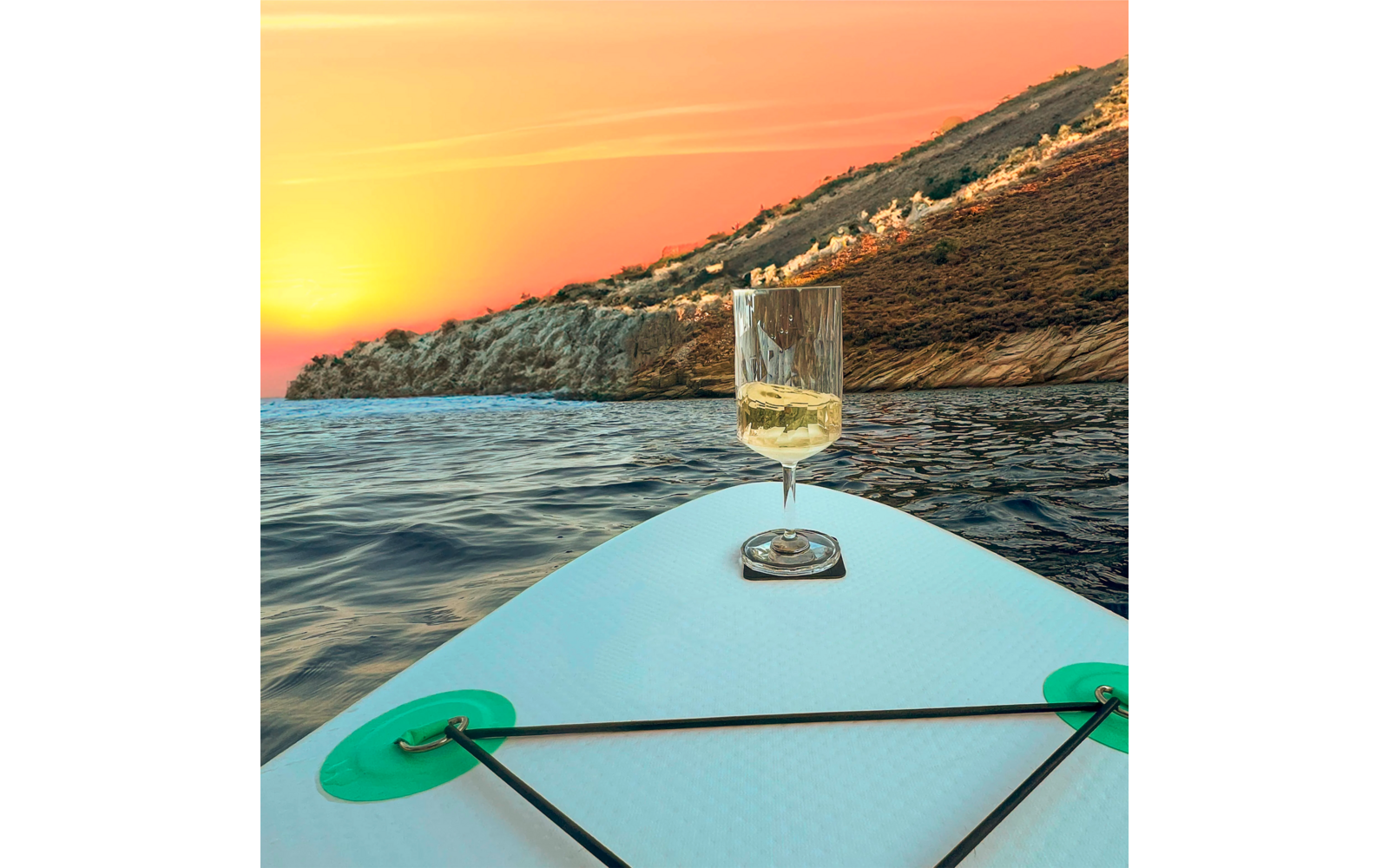 Wine glass on a stand-up paddleboard with a rocky shore and sunset in the background