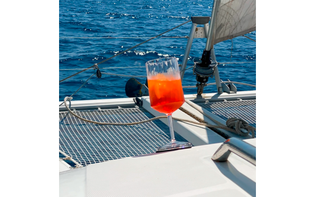 Glass with red drink on the netting of a sailboat, blue water surface in the background