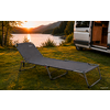 Cama de camping plegable sobre césped junto a la orilla de un lago, una furgoneta camper con la puerta abierta y montañas al fondo con el sol bajo en el horizonte