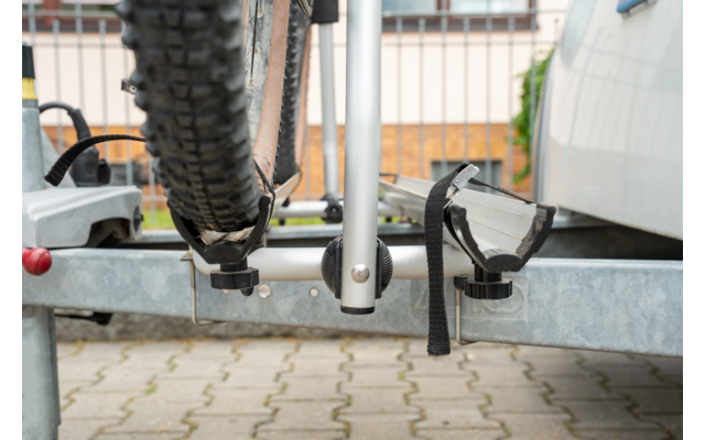 Primo piano di una gomma di bicicletta in un portabici metallico su un rimorchio, con cinghie di gomma e morsetti metallici; pavimentazione e parte di un veicolo sullo sfondo