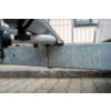 Primo piano di una traversa metallica e una staffa a U sotto un veicolo, scritta impressa sulla traversa, pavimentazione e facciata di un edificio sullo sfondo