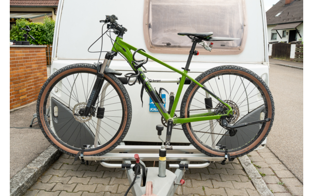 Mountain bike verde sul portapacchi posteriore di una roulotte bianca, gancio di traino e bordo stradale lastricato visibili