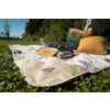 Couverture de pique-nique sur l'herbe avec le logo 'Happy Camp' visible, thermos, plateau en bois, une personne tient un livre vert, un coussin jaune et des baskets blanches visibles