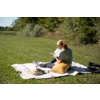 Personne assise sur une couverture dans un champ d'herbe, vue de dos, lisant un livre ; coussin jaune, bouteille et sac à côté de la personne ; arbres en arrière-plan