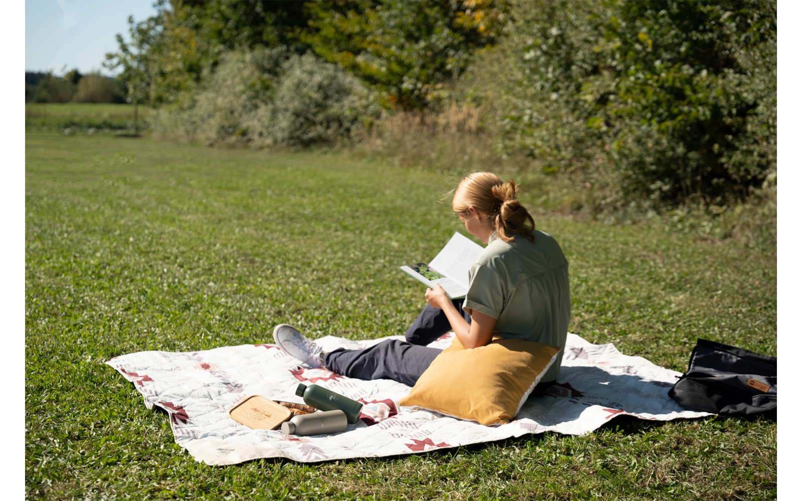 Personne assise sur une couverture dans un champ d'herbe, vue de dos, lisant un livre ; coussin jaune, bouteille et sac à côté de la personne ; arbres en arrière-plan