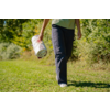 Bas du corps d'une personne portant un pantalon foncé et des chaussures blanches, marchant sur de l'herbe, tenant dans une main un tapis blanc roulé avec un motif, arbres et buissons en arrière-plan