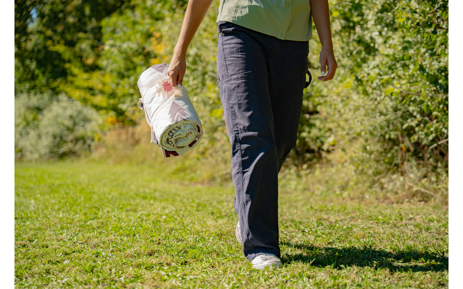 Bas du corps d'une personne portant un pantalon foncé et des chaussures blanches, marchant sur de l'herbe, tenant dans une main un tapis blanc roulé avec un motif, arbres et buissons en arrière-plan