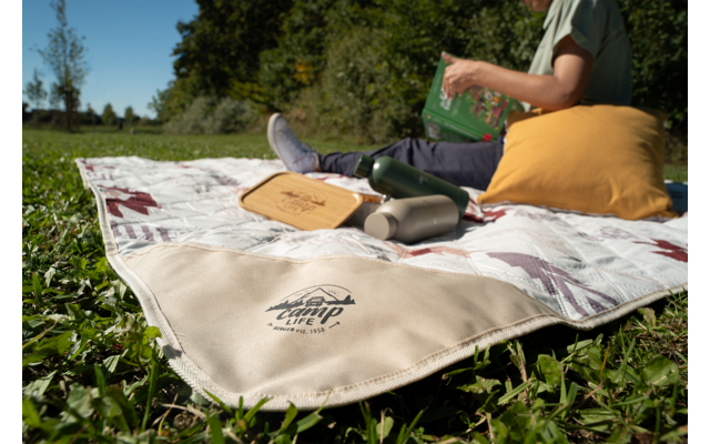 Couverture de pique-nique sur l'herbe avec le logo 'Happy Camp' visible, thermos, plateau en bois, une personne tient un livre vert, un coussin jaune et des baskets blanches visibles