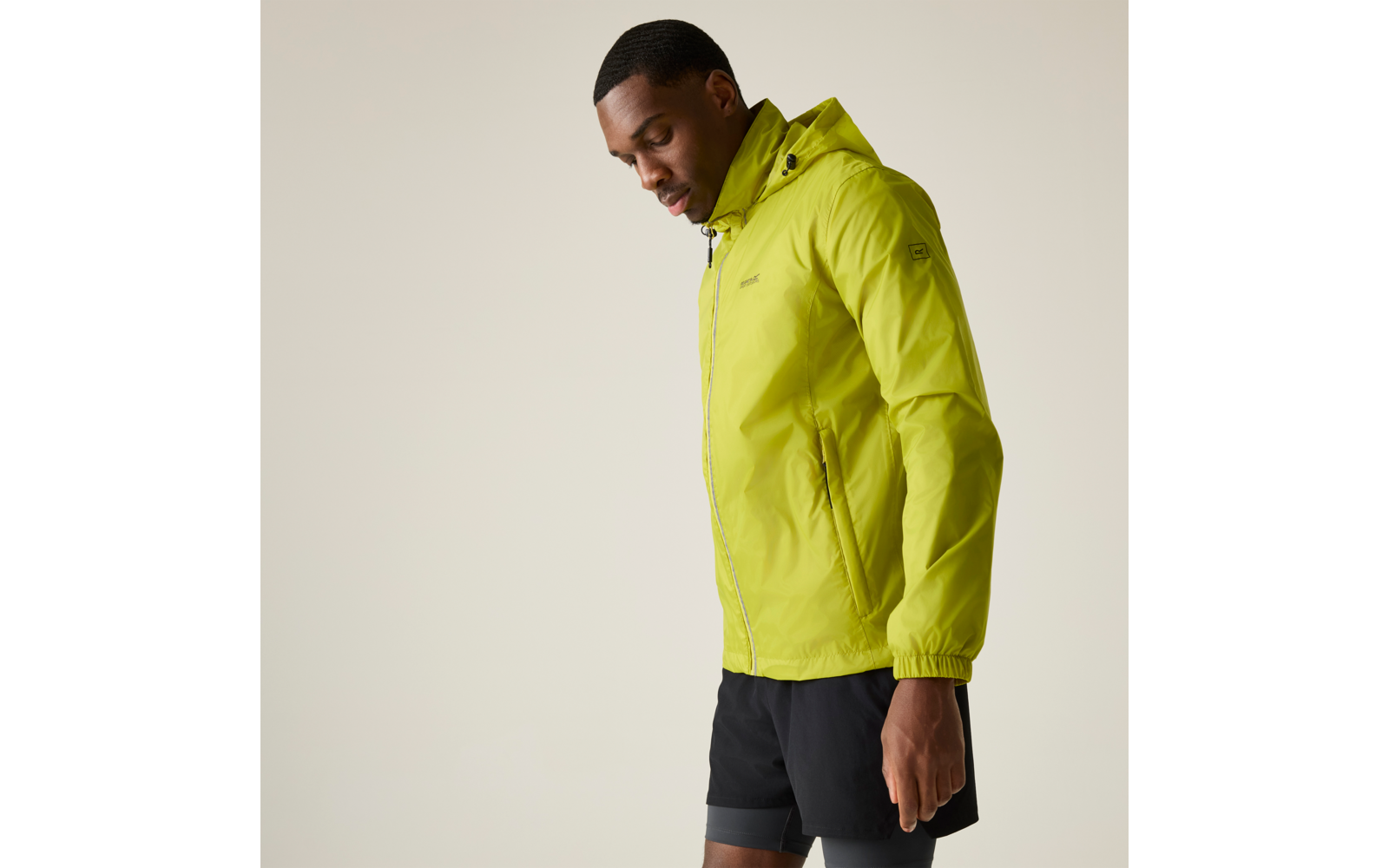 Seitlich stehender Mann in hellgrüner Kapuzenjacke und schwarzen Shorts vor hellem Hintergrund, Blick nach unten