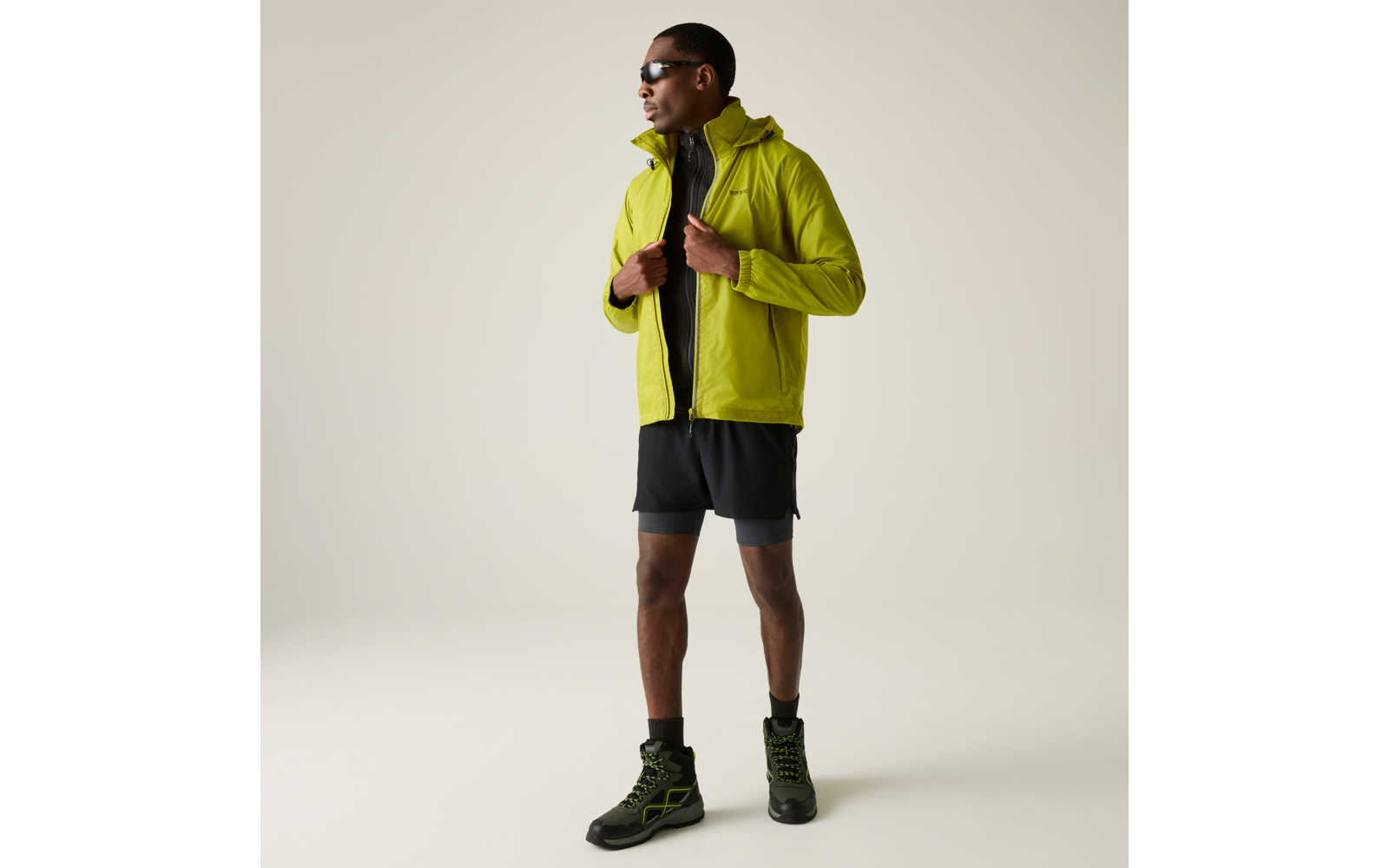 Person steht vor hellem Hintergrund, trägt leuchtend grüne Kapuzenjacke, schwarze Shorts mit Kompressionsschicht, schwarze hohe Sportschuhe mit grünen Details und Sonnenbrille; Hände halten den Reißverschluss der Jacke, Blick zur Seite