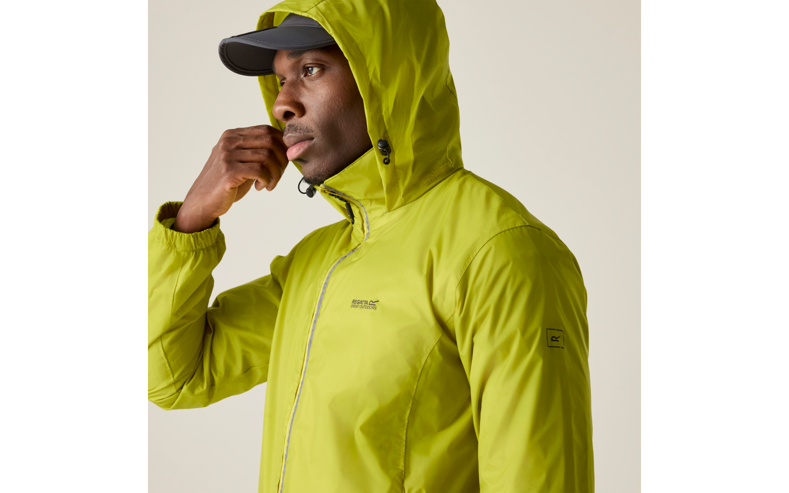 Person trägt eine limettengrüne Kapuzenjacke und eine schwarze Kappe, blickt nach links, Hand nahe am Gesicht, beiger Hintergrund