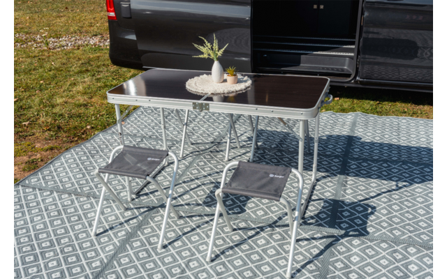 Opvouwbare tafel met donker blad en twee grijze opvouwbare krukjes op een patroon buitenkleed naast een open donkere bestelwagen; op tafel een rond dienblad met twee kleine vaasjes en een plant