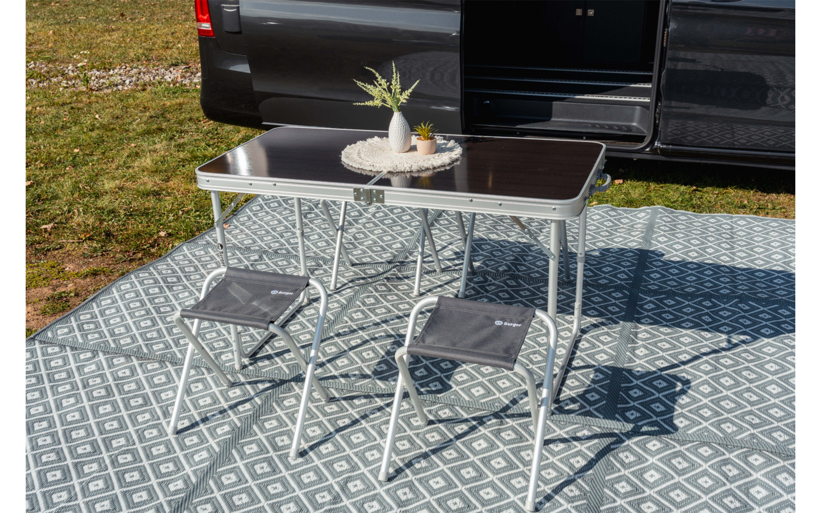 Opvouwbare tafel met donker blad en twee grijze opvouwbare krukjes op een patroon buitenkleed naast een open donkere bestelwagen; op tafel een rond dienblad met twee kleine vaasjes en een plant