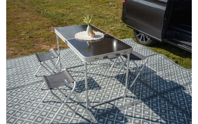 Opvouwbare metalen tafel met zwart blad op een gemêleerd buitenkleed, twee grijze opvouwbare krukjes, een ananas op een wit kanten kleedje op de tafel, geopende voertuigdeur met zichtbaar wiel, gras op de achtergrond