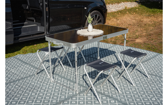 Opvouwbare tafel met klein vaasje en plant erop, vier opvouwbare krukjes op een patroonmat naast een open zwarte bus; gras en grind op de achtergrond