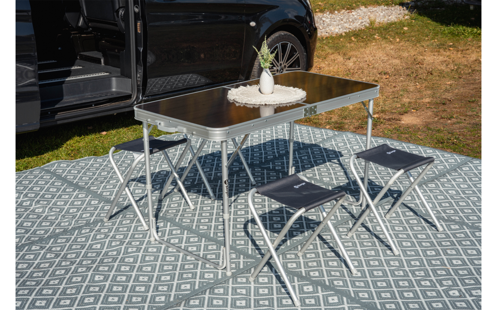 Opvouwbare tafel met klein vaasje en plant erop, vier opvouwbare krukjes op een patroonmat naast een open zwarte bus; gras en grind op de achtergrond