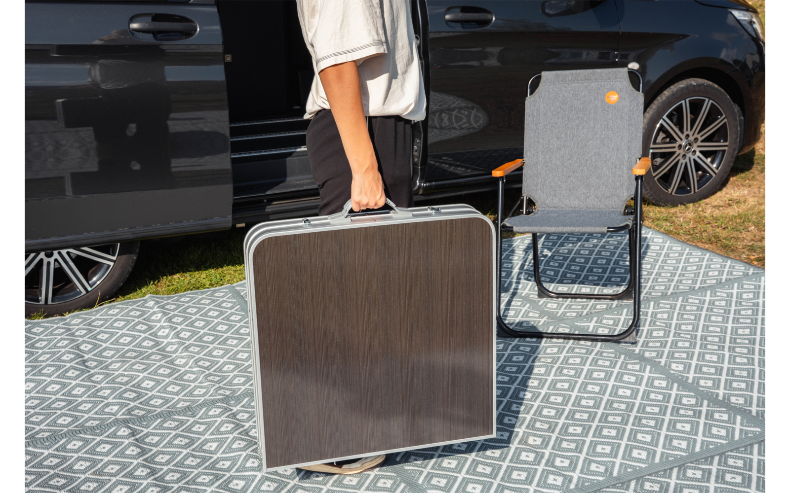 Gedeeltelijk zichtbare staande persoon in een wit T-shirt houdt een rechthoekige donkere draagkoffer met metalen frame; daarnaast een grijze vouwstoel met oranje details op een patroonmat; op de achtergrond een zwarte bus met open schuifdeur, gras aan de rand