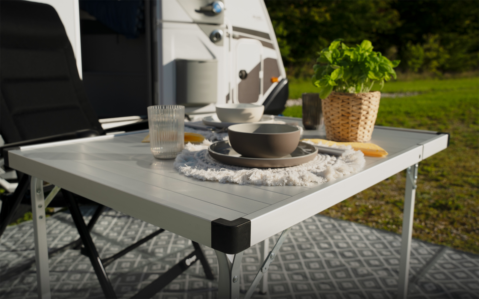 Weißer Klapptisch vor einem Wohnmobil mit einer Schale auf rundem Stoffuntersetzer, Trinkglas, Besteck, geflochtenem Pflanzkorb mit grünen Kräutern, Schneidebrett mit Brot, schwarzem Klappstuhl und gemustertem Bodenbelag