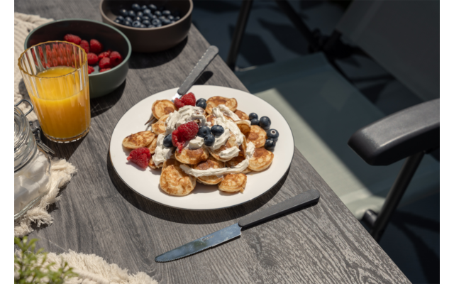 Witte schaal met kleine pannenkoekjes met room, frambozen en bosbessen op een houten tafel; daarnaast een glas sinaasappelsap, twee schaaltjes met bessen, mes en vork, een glazen pot, een textiel met franjes en een stoelarm in zonlicht
