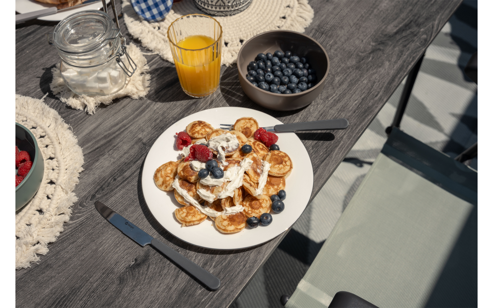 Bord met kleine pannenkoekjes, room, siroop, frambozen en bosbessen; vork op het bord, mes ernaast; glas sinaasappelsap, kom met bosbessen en glazen pot met suiker op een houten tafel; lichtgroene stoelrug zichtbaar