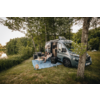 Grey camper van with open sliding door on grass, two people on folding chairs on a blue blanket, boxes and a lantern, trees and a lake in the background