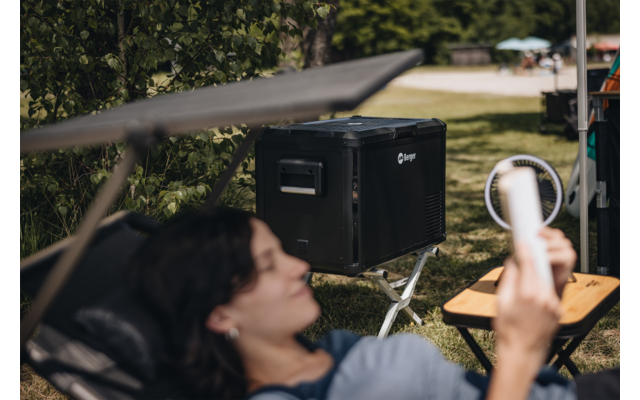 Person reclining in a lounge chair holding a smartphone; large black device on a stand, ring light and folding table in a park